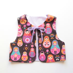 May include: A child's vest with a brown background and a repeating pattern of colorful Matryoshka dolls. The vest has a lavender ribbon closure and a white lining. The dolls are in shades of pink, orange, blue, and purple.