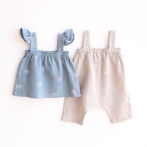 Può includere: Un completo per neonati composto da un top azzurro chiaro con spalline a volant e pantaloni beige. Entrambi i capi sono realizzati in tessuto strutturato e presentano un delicato motivo ricamato. Adatto a neonati e bambini piccoli.