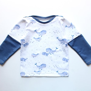 May include: A long-sleeved shirt featuring a white body with a pattern of blue whales and gray dots. The sleeves are a solid navy blue. The shirt has a crew neck and is likely for a child.
