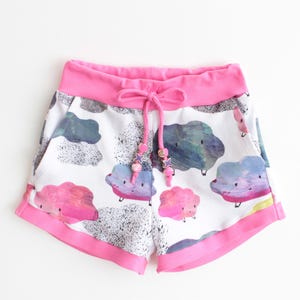 May include: White shorts with a pink waistband and trim, featuring a cloud pattern in shades of pink, blue, and gray. The shorts have a drawstring with decorative beads.