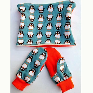 May include: A baby accessory set featuring a teal blue fabric with a repeating penguin pattern. The set includes a neck warmer and mittens with bright orange cuffs and lining. The penguin design has white bellies and black heads.