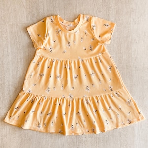 以下が含まれることがあります： イエローの半袖ワンピース。ティアードスカートと丸いネックラインが特徴。小さな白と黒の花柄模様。生地はリブ編みです。