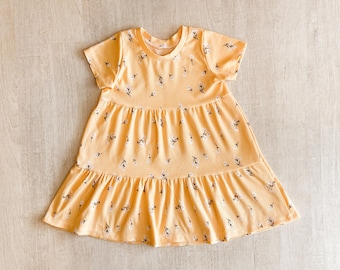 May include: A yellow, short-sleeved dress with a tiered skirt and a round neckline. The dress is patterned with small white and black floral sprigs. The fabric has a ribbed texture.