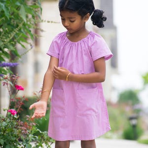 May include: A light purple, short-sleeved dress with two front pockets. The dress has a gathered neckline and a subtle checkered pattern. The girl is wearing a gold bracelet on each wrist. The dress is knee-length.