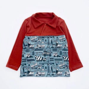 Op de afbeelding: Een shirt met lange mouwen met een baksteenrode kraag en mouwen. De borst heeft een blauw paneel met een herhaald patroon van bouwmachines, waaronder graafmachines, vrachtwagens en betonmixers. Het shirt is ontworpen voor kinderen.