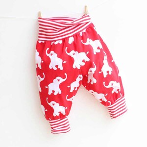 May include: Red baby trousers featuring a white elephant pattern. The waistband and cuffs have red and white stripes. The trousers are suspended on a washing line with a wooden peg.
