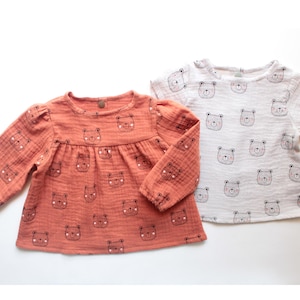 Könnte beinhalten: Zwei Baby-Shirts. Eines hat lange Ärmel in Korallenfarbe, das andere kurze Ärmel und ist weiß. Beide Shirts haben ein sich wiederholendes Muster von Bärengesichtern in schwarzer Umrandung mit rosa Akzenten.