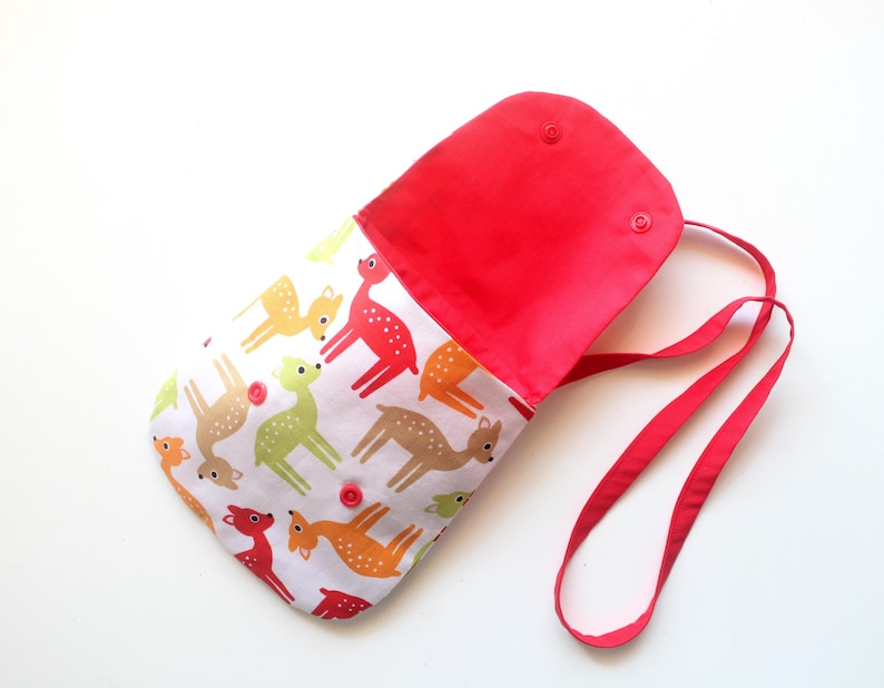 Puede incluir: Un bolso de hombro rojo y blanco con cierre de solapa. El bolso presenta un estampado de ciervos coloridos sobre un fondo blanco. El interior y la correa son rojos. El bolso tiene dos cierres a presi&oacute;n y una correa larga.