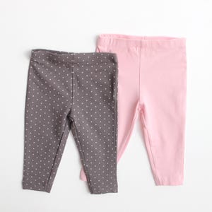 May include: Two pairs of baby leggings. One pair is solid light pink, and the other is dark gray with a white polka dot pattern. The leggings are made of a soft, stretchy material and have an elastic waistband.