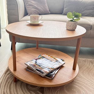 May include: A round, two-tiered wooden coffee table with a light brown finish. The top tier holds a small potted plant and a teacup on a saucer. The lower tier displays a stack of magazines. The table is in a living room setting.