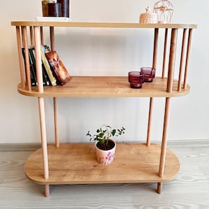 Puede incluir: Estantería de madera clara de tres niveles, con bordes redondeados y soportes verticales. La balda superior contiene velas y un plato pequeño. La balda central muestra libros y dos vasos morados. Una planta en maceta se encuentra en la balda inferior.