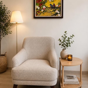 May include: A beige upholstered armchair with dark brown wooden legs, positioned next to a light wood semi-circular side table. The table holds a ribbed vase with green foliage and a small candle. A framed painting of birds hangs above.