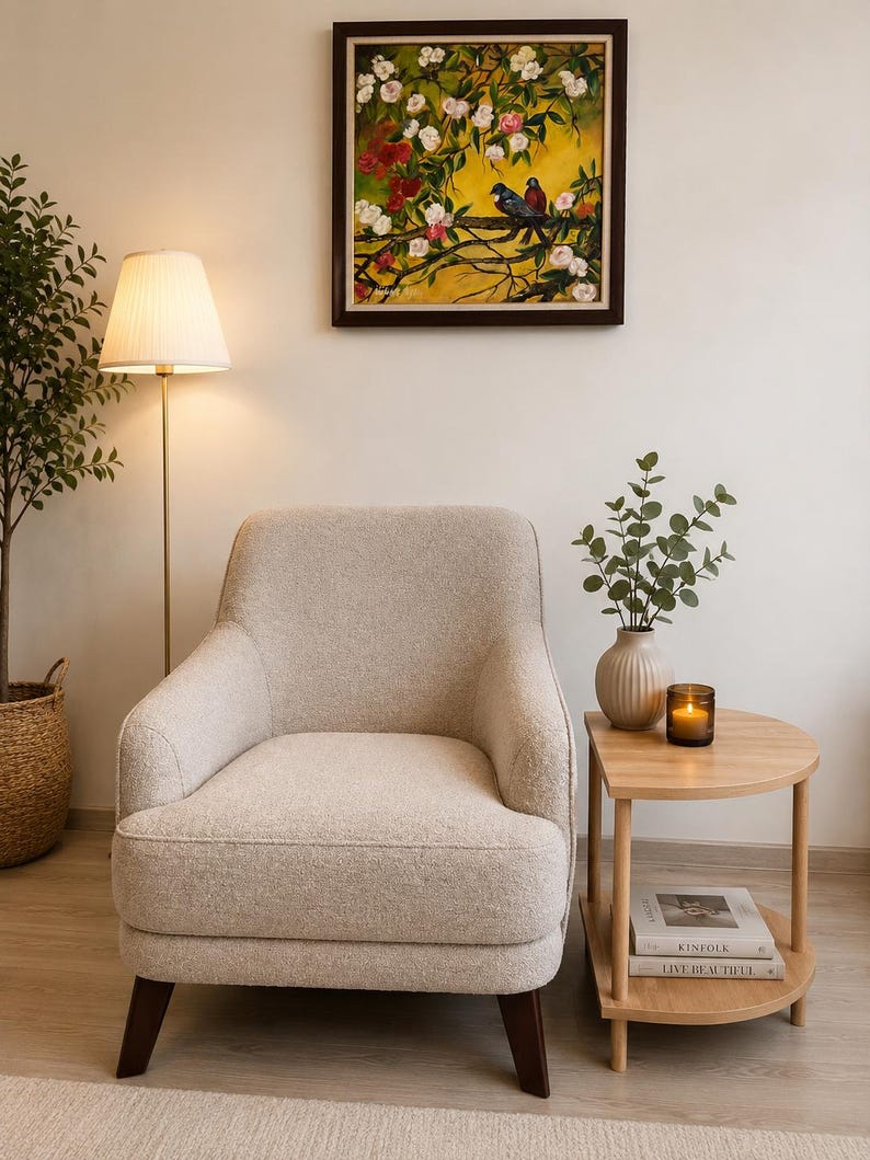 May include: A beige upholstered armchair with dark brown wooden legs, positioned next to a light wood semi-circular side table. The table holds a ribbed vase with green foliage and a small candle. A framed painting of birds hangs above.