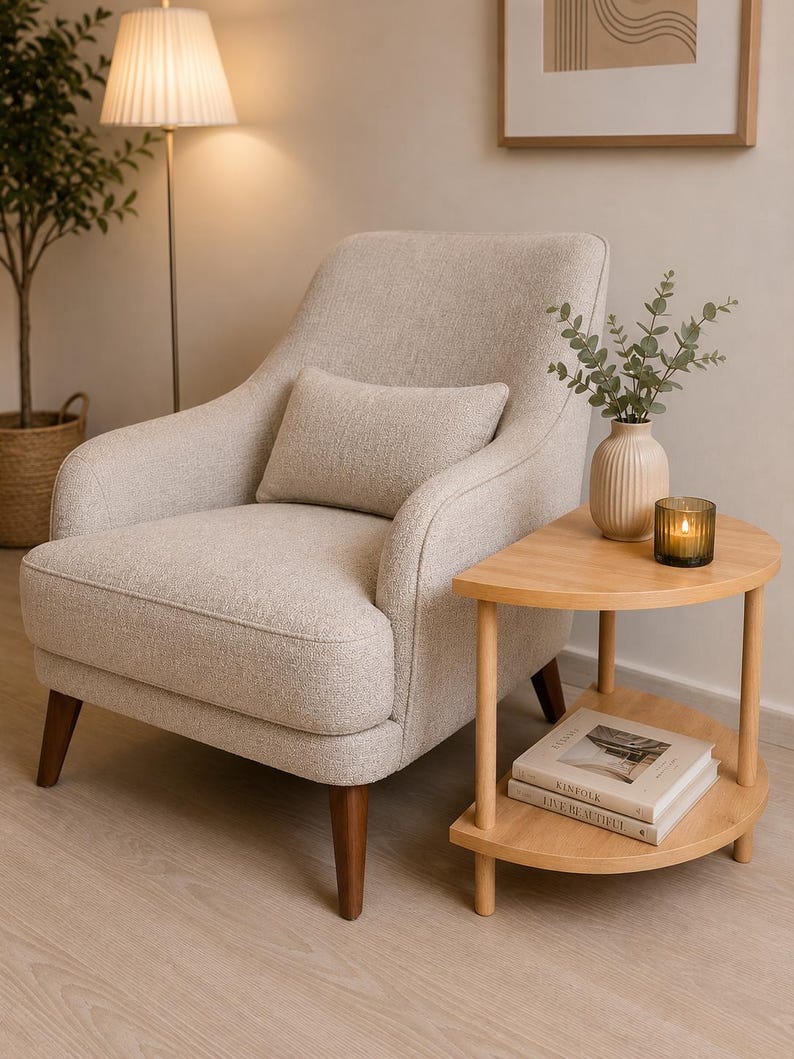 May include: An upholstered armchair in a light beige textured fabric with a matching rectangular lumbar pillow, supported by four dark brown tapered wooden legs, positioned next to a two-tiered semi-circular light wood side table.