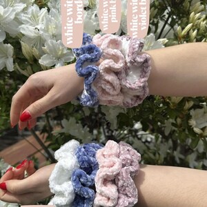 Four scrunchies in different colors. The scrunchies are made of a soft, fluffy material. The colors are white, blue, baby pink, and dusty violet. The scrunchies are stacked on top of each other. The scrunchies are labeled with the words 'white', 'blue', 'baby pink', and 'dusty violet'.