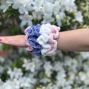 four scrunchies in violet, pink, white, and blue velvet. The scrunchies are stacked on top of each other.