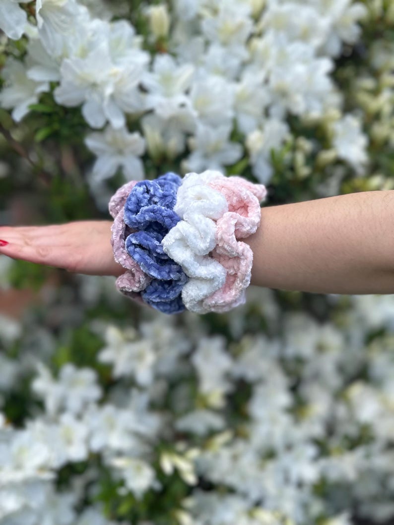 four scrunchies in violet, pink, white, and blue velvet. The scrunchies are stacked on top of each other.