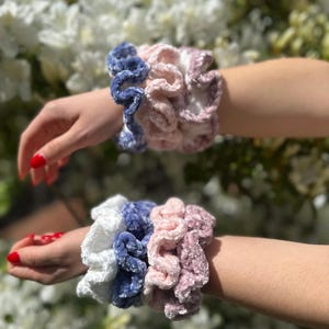 : Four scrunchies in white, blue, pink and a darker pink color. The scrunchies are made of a soft, fuzzy material and are stacked on top of each other.