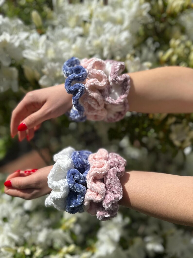: Four scrunchies in white, blue, pink and a darker pink color. The scrunchies are made of a soft, fuzzy material and are stacked on top of each other.