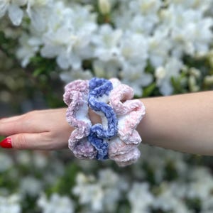 3 scrunchies made of soft, fuzzy yarn. One scrunchie is a dusty violet with white, one is light pink with white,  and the other is a light blue with white.