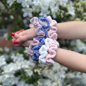2 hands clasped together featuring three velvet crochet scrunchies on each wrist in pink, white, and blue velvet fabric.
