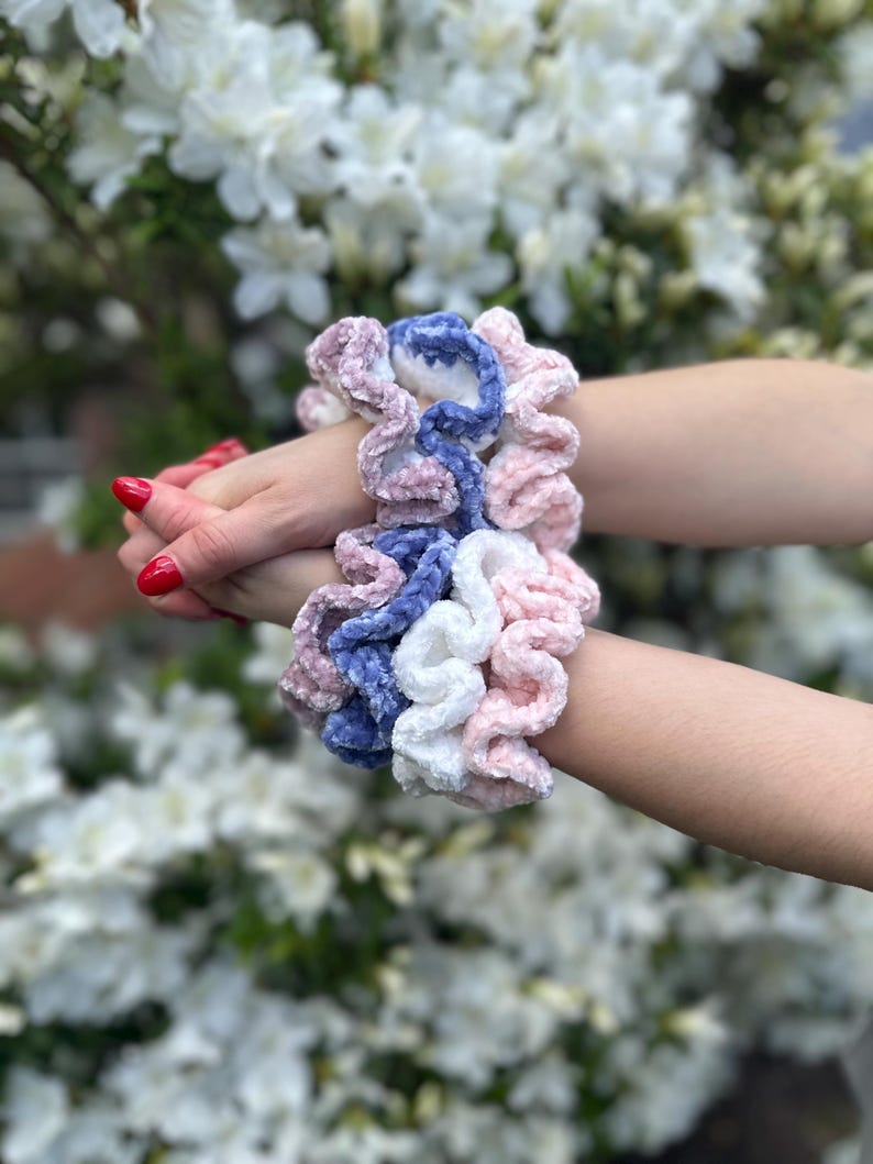 2 hands clasped together featuring three velvet crochet scrunchies on each wrist in pink, white, and blue velvet fabric.