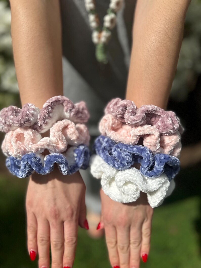 Three sets of colorful crocheted hair scrunchies. The scrunchies are layered on top of each other. The bottom layer is blue, the middle layer is white, and the top layer is pink.