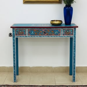 Può includere: Uno specchio con cornice dorata e parte superiore ad arco è appeso sopra una consolle turchese. Il tavolo ha un cassetto ed è decorato con motivi floreali colorati. Un vaso blu con fiori e una ciotola dorata sono sul tavolo.