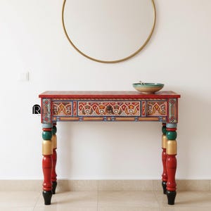 Può includere: Una consolle dipinta a mano, rossa, con motivi intricati blu, gialli e verdi. Il tavolo ha un singolo cassetto ed è sostenuto da quattro gambe con un design a blocchi di colore. Uno specchio rotondo dorato è appeso sopra il tavolo.