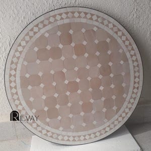 May include: Round mosaic table with a geometric pattern in shades of pink and white. The table has a black border and a decorative edge with diamond shapes. The mosaic tiles create a visually appealing surface.