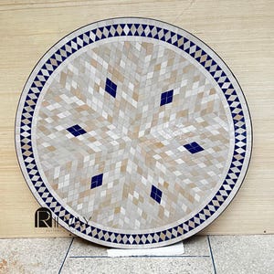 May include: Circular mosaic table top with a geometric design. The centre has a starburst pattern in beige, white, and navy blue. The edge is navy blue with a repeating diamond pattern. Suitable for indoor or outdoor use.
