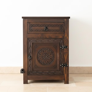Może przedstawiać: Ciemnobrązowa drewniana szafka nocna ze skomplikowanymi rzeźbionymi wzorami kwiatowymi i geometrycznymi. Posiada szufladę z okrągłym uchwytem i drzwiczki szafki z centralnym okrągłym motywem. Szafka nocna ma prostokątny kształt i stoi na czterech krótkich nogach.