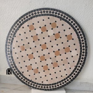 May include: Round mosaic tile table with a beige, brown, and black geometric pattern. The table has a black border with a diamond pattern. The center of the table features a repeating pattern of beige, brown, and black tiles.