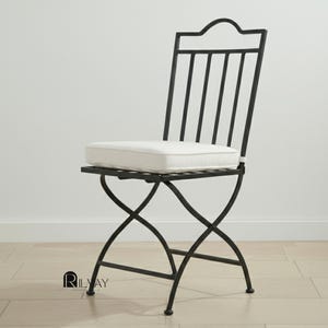 May include: Black metal outdoor chair with a white cushion. The chair has a decorative back with vertical bars and a crossed leg design. The cushion is rectangular and appears to be made of a soft fabric. The chair is designed for outdoor use.