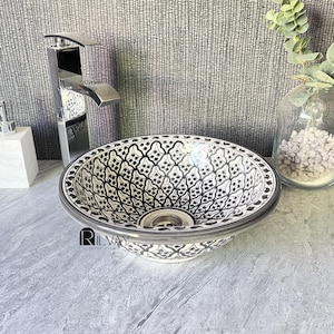 Puede incluir: Un lavabo redondo de cerámica con un patrón en blanco y negro y borde plateado. El lavabo está sobre una encimera gris. Se ve un grifo cromado y un dispensador de jabón. El fondo es una pared gris texturizada.