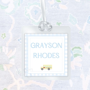 May include: A clear, square luggage tag with a light blue gingham border. The tag features the name "GRAYSON RHODES" in light blue text and a small yellow school bus illustration. The tag is attached to a clear loop.