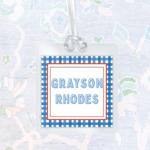 May include: A square luggage tag featuring a blue and white gingham border, a thin red inner frame, and the name GRAYSON RHODES printed in light blue capital letters on a white background, attached to a clear plastic loop for secure fastening.