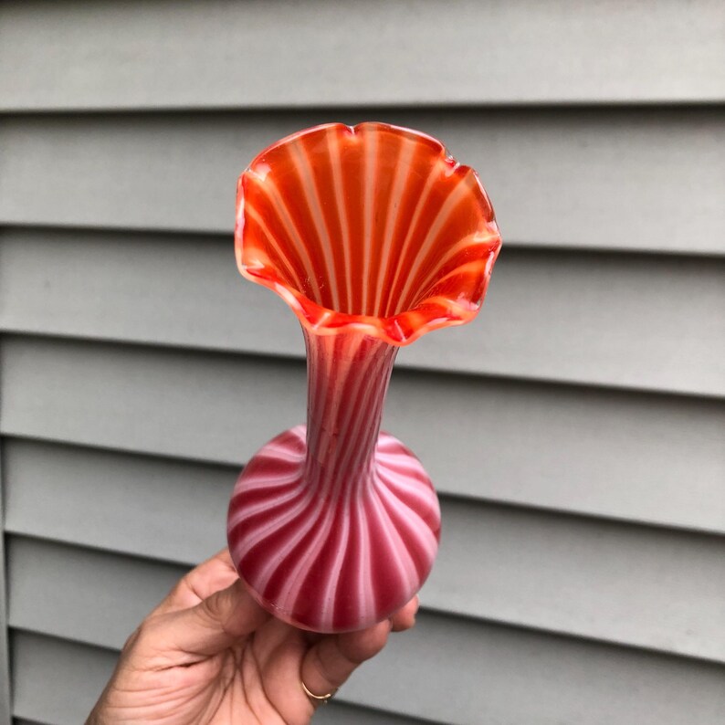 May include: A vintage glass vase with a flared, ruffled rim. The vase features a vibrant color scheme of red and white stripes, creating a striking visual effect. The base is round, and the neck is slender, leading to the open, flower-like top.