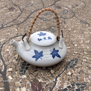 May include: A small white ceramic teapot with a brown handle and blue floral designs. The teapot has a lid with Chinese characters that read "Tea Pot".