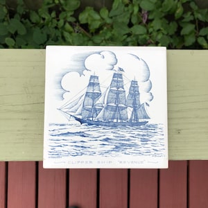 May include: A blue and white ceramic tile featuring a drawing of a clipper ship sailing on the sea. The tile is inscribed with the words "Clipper Ship 'Revenue'".