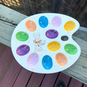 May include: White ceramic Easter egg holder shaped like a painter's palette with 12 individual egg-shaped depressions. The palette features a colorful bunny holding a paintbrush and palette.
