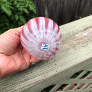 May include: A small, round glass vase with alternating red and white stripes. The vase has a clear base and a small blue and white label that reads "WAN". The vase is held in a hand.