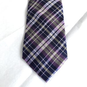 May include: A necktie featuring a diagonal plaid pattern in shades of dark navy blue, muted olive green, and soft lavender, accented by thin white intersecting lines that create a structured grid across the entire surface of the fabric.
