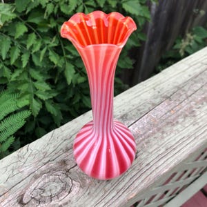 May include: A vintage glass vase with a ruffled rim and a striped pattern in shades of red and white. The vase has a slender neck and a rounded base. The vase is approximately 8 inches tall.