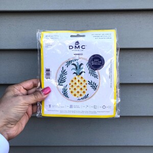 May include: A cross-stitch kit featuring a pineapple with green leaves in a wooden embroidery hoop. The kit is packaged in a clear plastic bag with a yellow and white label that says "DMC Mouline Special".