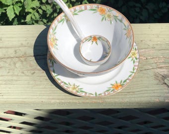 May include: A white porcelain soup bowl with a floral design in shades of yellow, orange, and green. The bowl is nested inside a matching saucer. A white porcelain ladle with a floral design is resting inside the bowl.