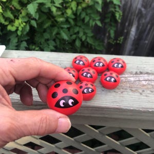 May include: A collection of red wooden ladybug-shaped toys with black spots and cartoonish eyes. One is held in a hand, with others scattered on a wooden surface. The toys are a vibrant red with black details.