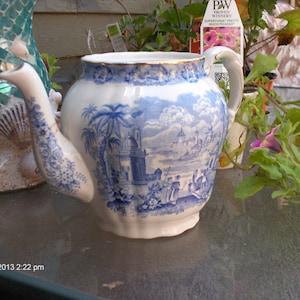 May include: A blue and white porcelain teapot with a floral design and a scene of a town with palm trees. The teapot has a spout and a handle. The teapot is sitting on a table with a green plant in the background.