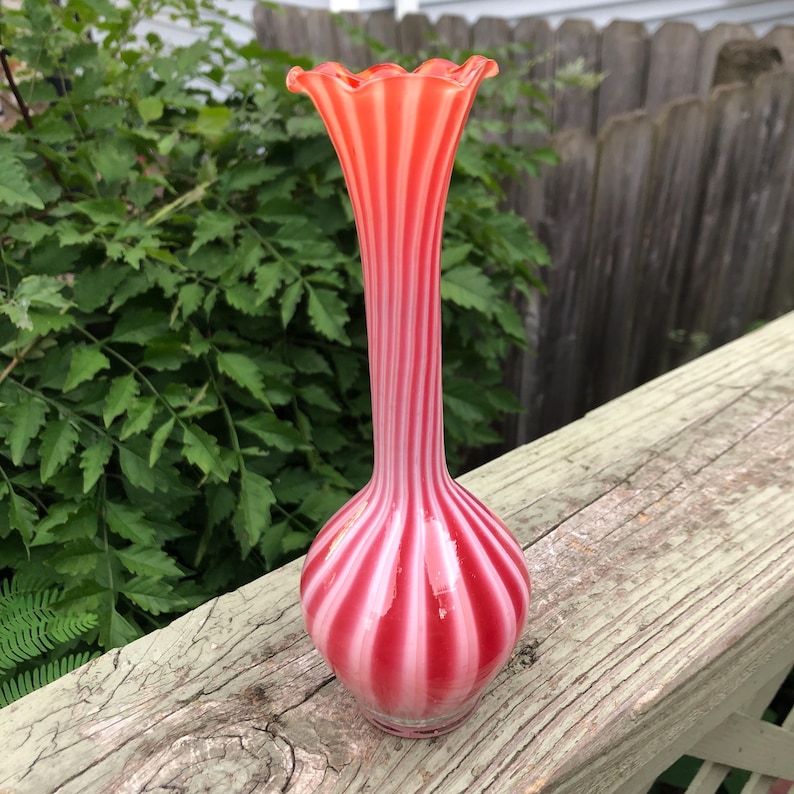 May include: A tall, slender glass vase with a ruffled rim. The vase features alternating vertical stripes of red and white. The vase is approximately 10 inches tall and has a bulbous base.