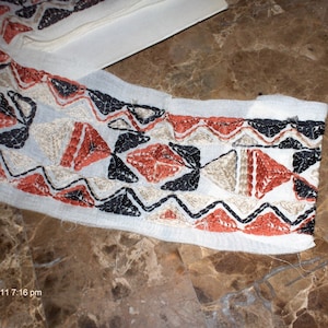 May include: White fabric with a geometric pattern embroidered in black, red, and beige thread. The pattern is a series of triangles and zigzags.